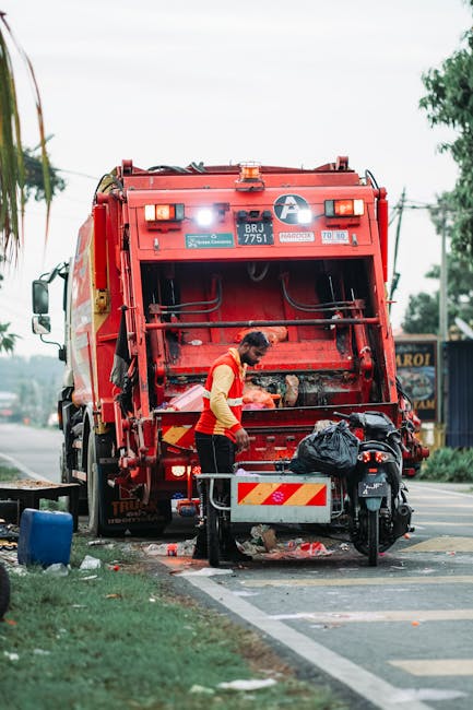 A worker dressed in a red and yellow uniform is operating a large red rubbish collection vehicle on a street, standing at the rear loading area where the hatch is open. The vehicle's back panel features reflective red and yellow striped markings, and various equipment and hoses are attached to the exterior. The worker appears to be handling black waste bags or debris, which are seen on the ground near the vehicle. The scene is set outdoors during daytime, with a nearby sidewalk, a patch of grass, and a few trees visible in the background, along with utility poles and wires. The environment suggests an urban or suburban area where rubbish removal services, such as those offered by Rubbish Removal Colliers Wood, conduct on-site waste collection or clearance. The overall atmosphere emphasizes scheduled waste disposal or private rubbish collection, supporting the context of alternative refuse management methods outside of local authority collection services.