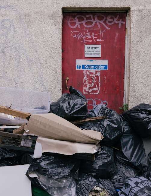 A collection of black plastic rubbish bags and discarded cardboard boxes piled in front of a red metal door embedded in an exterior beige concrete wall. The door features various graffiti tags and a white sign stating 'No Money' and 'Keep Clear' in blue and white. The rubbish bags vary in size and appear to contain household or waste items, with some partially torn or crumpled. There are additional loose pieces of cardboard and debris among the refuse, suggesting an on-site clear-out or informal rubbish disposal. The environment looks like an alleyway or service area, with limited natural light illuminating the scene and emphasizing the cluttered nature of the waste. This scene illustrates private rubbish handling options that may require independent collection, such as those offered by local waste removal services like Rubbish Removal Colliers Wood, to properly tidy and manage on-site waste and prevent unlawful dumping in communal or commercial settings.