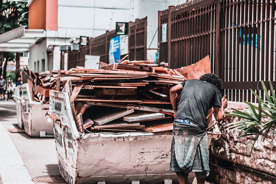A man with curly dark hair, wearing a black T-shirt and a plaid skirt, is standing next to a large white skip filled with a substantial pile of construction and demolition debris, including weathered terracotta roof tiles, wooden planks, and metal sheets. The debris appears to be stacked haphazardly, occupying most of the container and extending slightly over the edges. The surface of the skip shows signs of dirt, rust, and wear, indicative of heavy use. In the background, there is a high chain-link fence coated in rust, running parallel to the sidewalk, with some greenery and a building partially visible behind it. To the left of the skip, a few pedestrians can be seen walking along the pavement. Overhead, there are street signs and small surveillance cameras mounted on poles attached to the building, which features a modern exterior with clean lines and a neutral color palette. The lighting suggests natural daylight, with slight shadows cast on the scene, illustrating an urban environment where private waste removal or on-site clearance might be taking place, aligning with other alternative rubbish handling services provided by Rubbish Removal Colliers Wood.