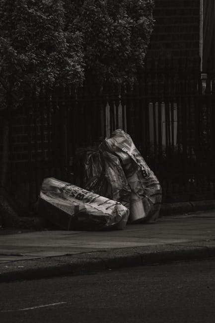 Three large black plastic rubbish bags, slightly crumpled and tied at the top, are positioned on the pavement near a black metal fence with vertical slats. The bags appear to contain waste materials, possibly household rubbish, and are placed closely together, leaning slightly against each other. In the background, dense foliage of leafy trees or shrubs extends upwards, partially obscuring the top of the fence. The setting is outdoor, on a street or residential area with a smooth asphalt road and a concrete sidewalk. The scene is captured in natural daylight, with shadows cast on the ground, emphasizing the textures of the bags and the surrounding environment. The overall scene exemplifies typical informal rubbish disposal, which may fall under private waste collection services like those offered by Rubbish Removal Colliers Wood, facilitating alternative waste handling outside regular municipal refuse collection.