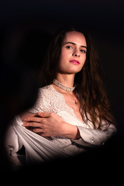 A woman with long, wavy brown hair is seated indoors against a dark background. She is wearing a white lace dress with intricate floral patterns and a high neckline, along with a layered pearl necklace. Her left arm is crossed over her chest, with her hand resting on her right shoulder, which is covered by a long-sleeved white shirt with a buttoned cuff. Her facial expression is neutral with slightly parted lips, and her gaze is directed towards the camera. The lighting highlights her face and upper body, creating a contrast with the dark surroundings. The composition and lighting suggest a professional portrait setting, with attention to detail in the clothing and accessories. The image appears unrelated to rubbish removal services or waste management, and solely depicts a portrait of a woman in elegant attire.