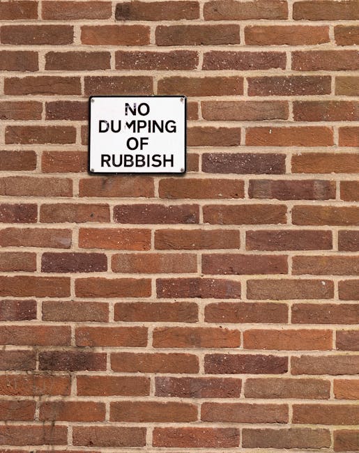 A rectangular metal sign mounted on a red brick wall, displaying the message 'NO DUMPING OF RUBBISH' in black uppercase letters on a white background. The sign is positioned centrally on the wall, which features a traditional running bond pattern with reddish-brown bricks and light-colored mortar joints. The wall's surface is uniform, with no other objects or markings visible. The lighting appears natural, with soft shadows cast onto the wall beneath the sign, suggesting daylight. This scene relates to local waste management regulations, highlighting an area where private rubbish removal services, such as those offered by Rubbish Removal Colliers Wood, might be necessary for proper waste disposal.