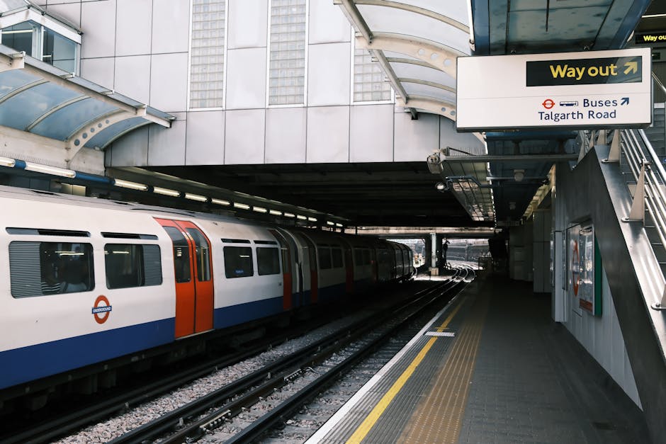 A modern train station platform with a partially covered, curved glass and metal roof structure supported by metal beams. On the left side of the image, a white and orange underground train is stationary on the tracks, with visible windows and the London Underground roundel logo near the front. The platform surface is made of textured grey paving with a yellow tactile strip along the edge for safety. In the background, the train tracks extend into a darker tunnel or underpass area. Above the platform, there is a prominent white sign with black and yellow text indicating the way out and directions to buses and Talgarth Road, mounted on the station's metallic framework. The environment is clean and well-maintained, suggesting the station is an integral part of urban public transportation. Rubbish removal services, such as those provided by Rubbish Removal Colliers Wood, often assist with waste management at such busy transport hubs, ensuring cleanliness and safety.
