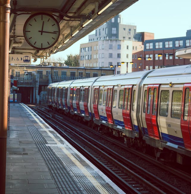 A city train with a silver exterior accented by red and blue stripes is stationed at an outdoor metropolitan railway platform. The train's windows reflect the surrounding urban environment, and the train cars are aligned parallel to the platform edges. The platform surface appears paved with rectangular tiles, and tactile paving strips are visible near the platform edge for safety. Overhead, a large round clock with black hands and a white face is mounted under a sheltered canopy, indicating the time as approximately 4:02. The shelter has exposed metal structural elements and linear lighting fixtures. In the background, a mixture of modern and older multi-storey buildings, including glass-fronted and brick facades, creates an urban skyline. The scene is illuminated by natural daylight, suggesting early evening or morning, with soft shadows cast on the platform, indicating clear weather. The image depicts a typical urban railway station environment, relevant to private or independent railway services and alternative transit options within a city context.