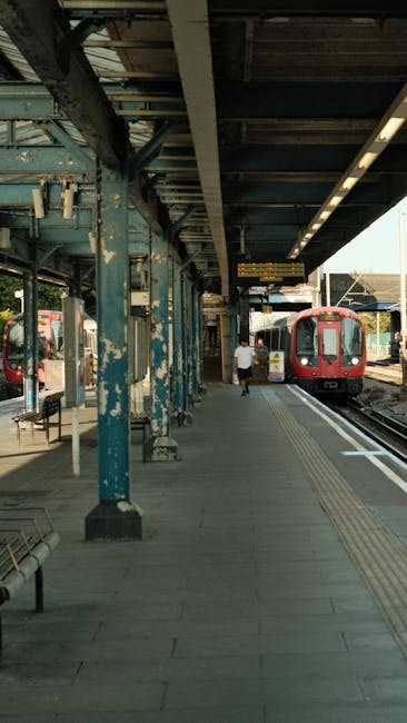 A modern train station platform with a partially covered, curved glass and metal roof structure supported by metal beams. On the left side of the image, a white and orange underground train is stationary on the tracks, with visible windows and the London Underground roundel logo near the front. The platform surface is made of textured grey paving with a yellow tactile strip along the edge for safety. In the background, the train tracks extend into a darker tunnel or underpass area. Above the platform, there is a prominent white sign with black and yellow text indicating the way out and directions to buses and Talgarth Road, mounted on the station's metallic framework. The environment is clean and well-maintained, suggesting the station is an integral part of urban public transportation. Rubbish removal services, such as those provided by Rubbish Removal Colliers Wood, often assist with waste management at such busy transport hubs, ensuring cleanliness and safety.